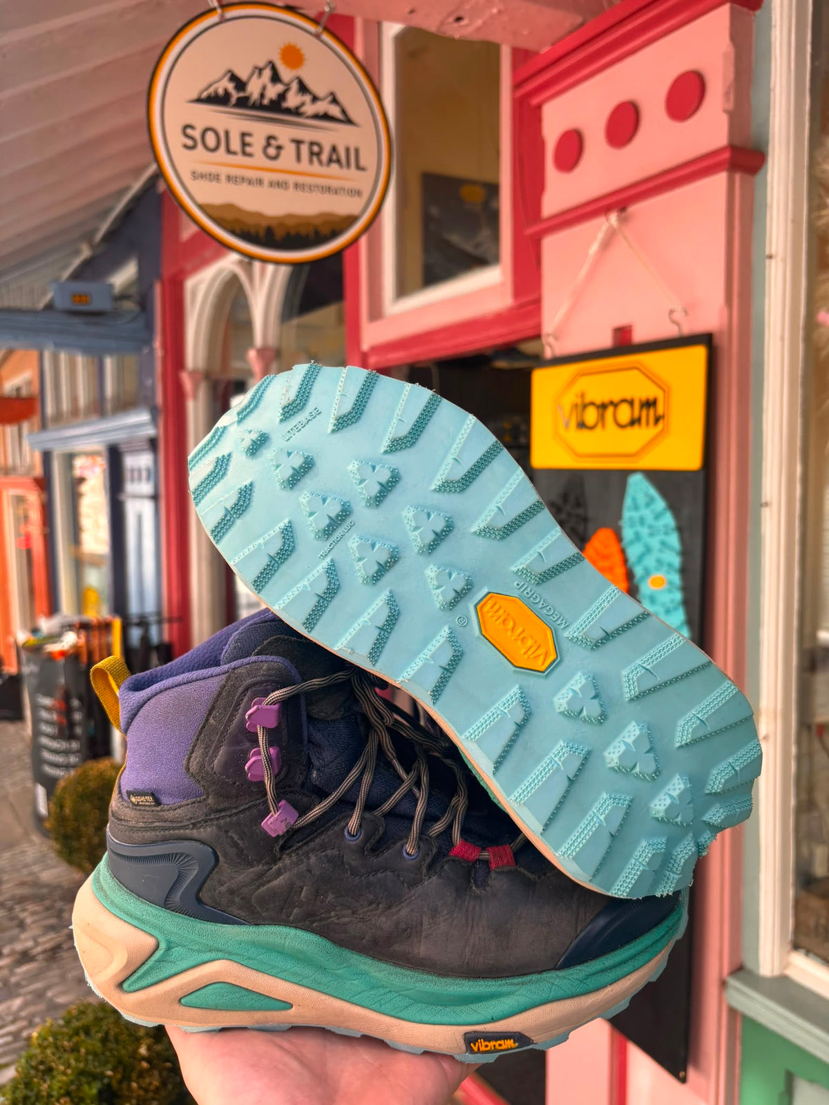 A hiking boot held up by a hand in front of a storefront, showing the outsole and the side of the boot. The boot has a dark gray and purple upper with a teal midsole and a yellow Vibram logo on the outsole. Vibram Resoling Trail