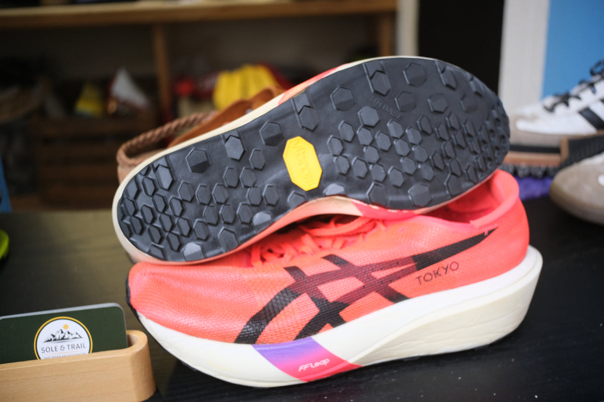 A pair of ASICS Gel-Kayano running shoes in a vibrant coral pink color, with one shoe stacked on top of the other to show the black Vibram sole and the white midsole. The ASICS logo is visible on the side, and the shoes are displayed on a dark tabletop with a blurred background. Running/Trail Resole - Sole and Trail