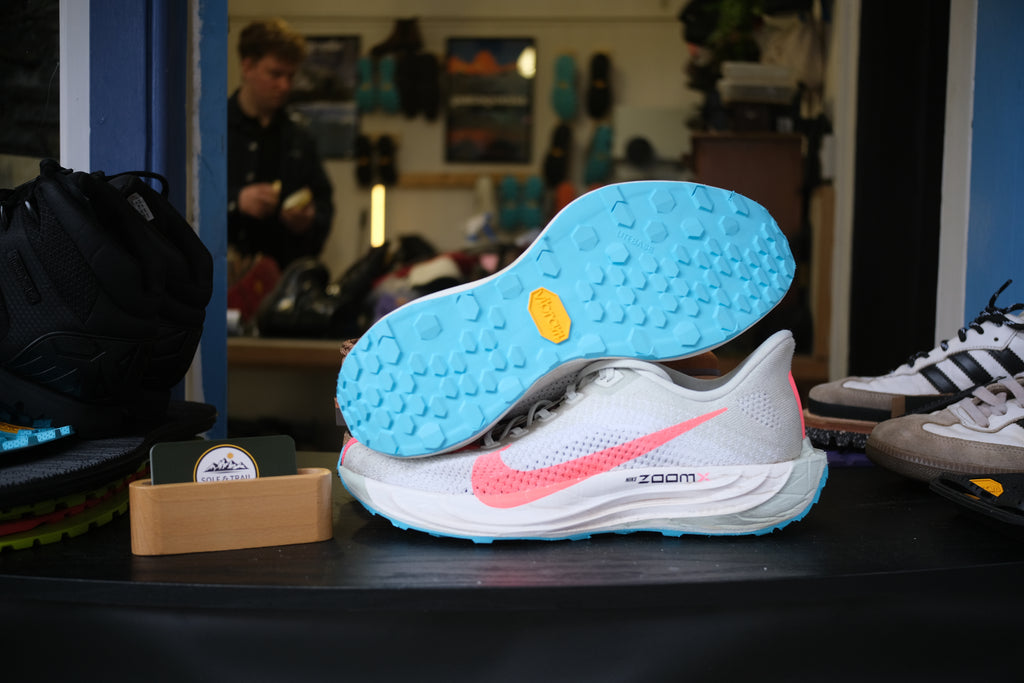 A white Nike running shoe with a bright pink Swoosh and a light blue Vibram sole, displayed on a dark table inside a shoe store. Vibram Speedster, Running/Trail Resole - Sole and Trail 