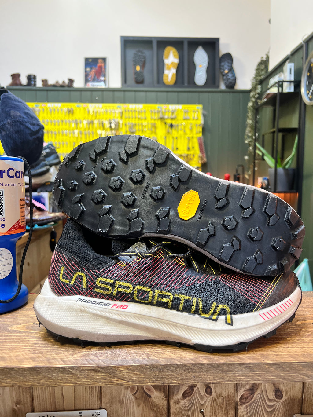 A pair of La Sportiva Pegasus PRO trail running shoes displayed on a wooden shelf in a retail store. One shoe is positioned upright to show the side profile, featuring a black and red patterned upper with yellow 'LA SPORTIVA' branding, a white midsole, and a black outsole. Running/Trail Resole - Sole and Trail
Vibram zegatarc