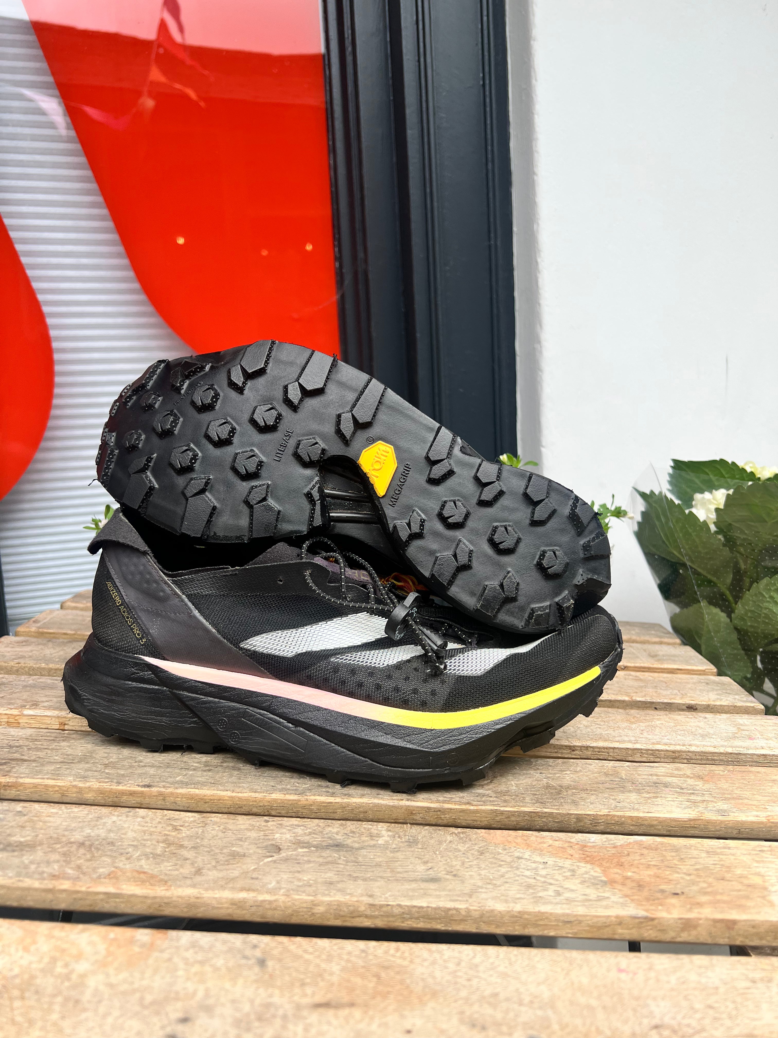 A pair of black trail running shoes with a yellow Vibram logo and a pink accent stripe, displayed on a wooden slatted surface with one shoe showing its treaded sole. Running/Trail Resole - Sole and Trail
Vibram zegatarc