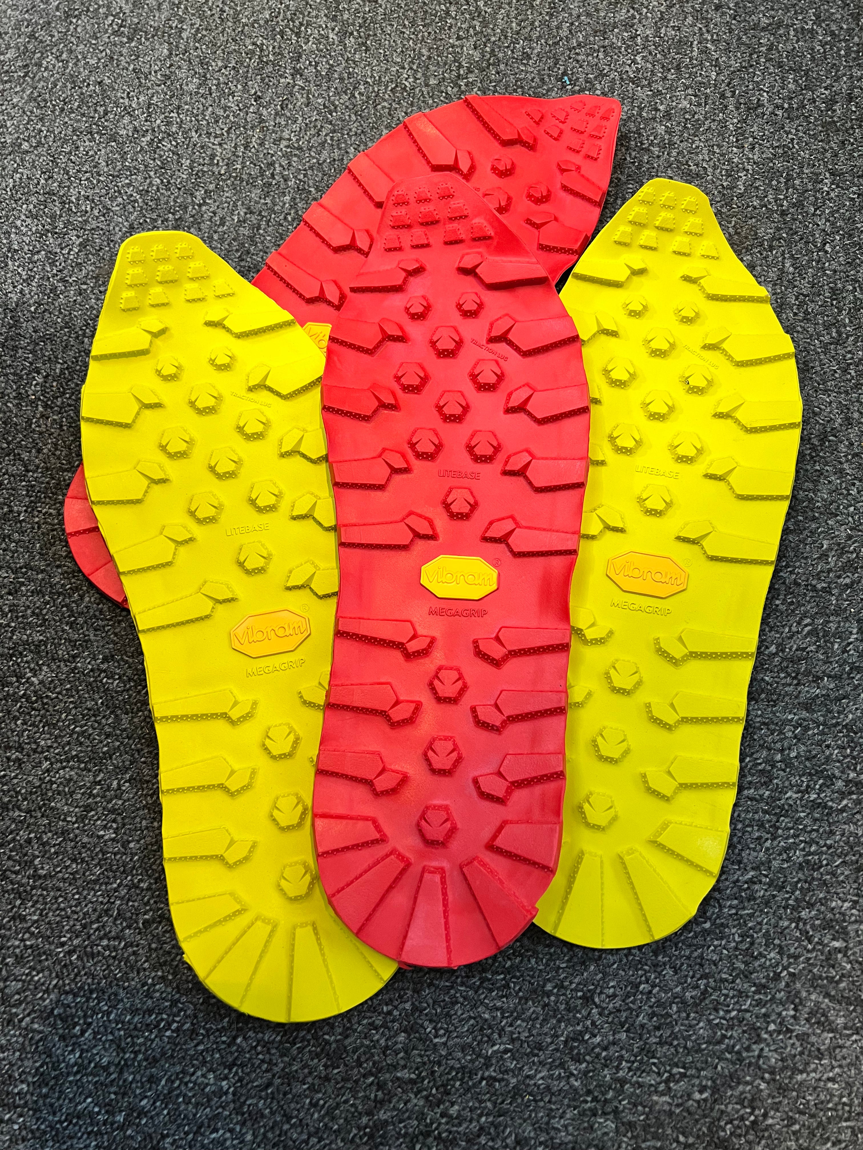 Three Vibram shoe insoles displayed flat on a dark gray carpeted surface, featuring a central red sole with a yellow Vibram logo and flanked by two yellow soles. Running/Trail Resole - Sole and Trail Vibram Zegatrac