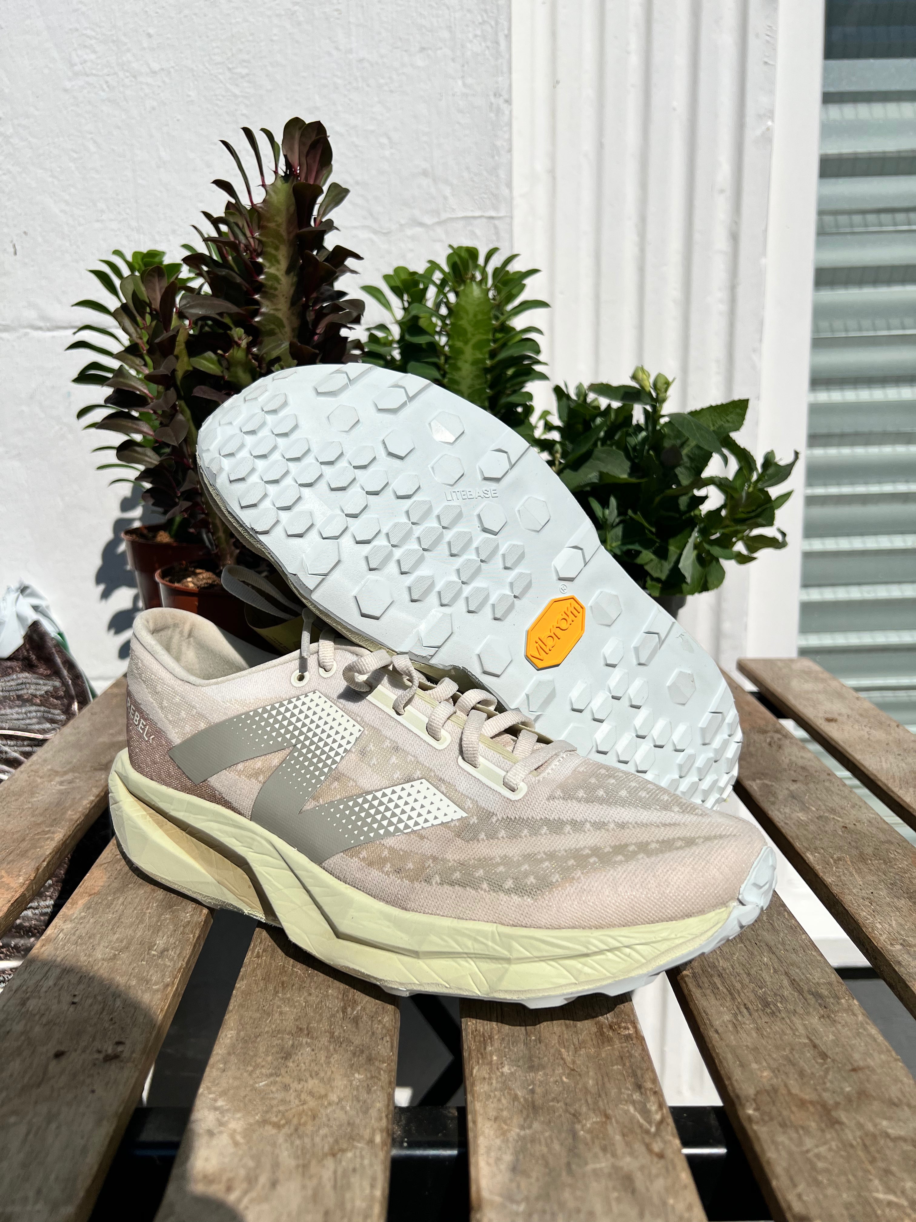 A pair of New Balance 1080v4 running shoes in a light beige and gray colorway, displayed on a wooden slatted bench with green plants in the background. One shoe is positioned upright while the other is turned to show the textured white outsole. Sole and Trail Kendal