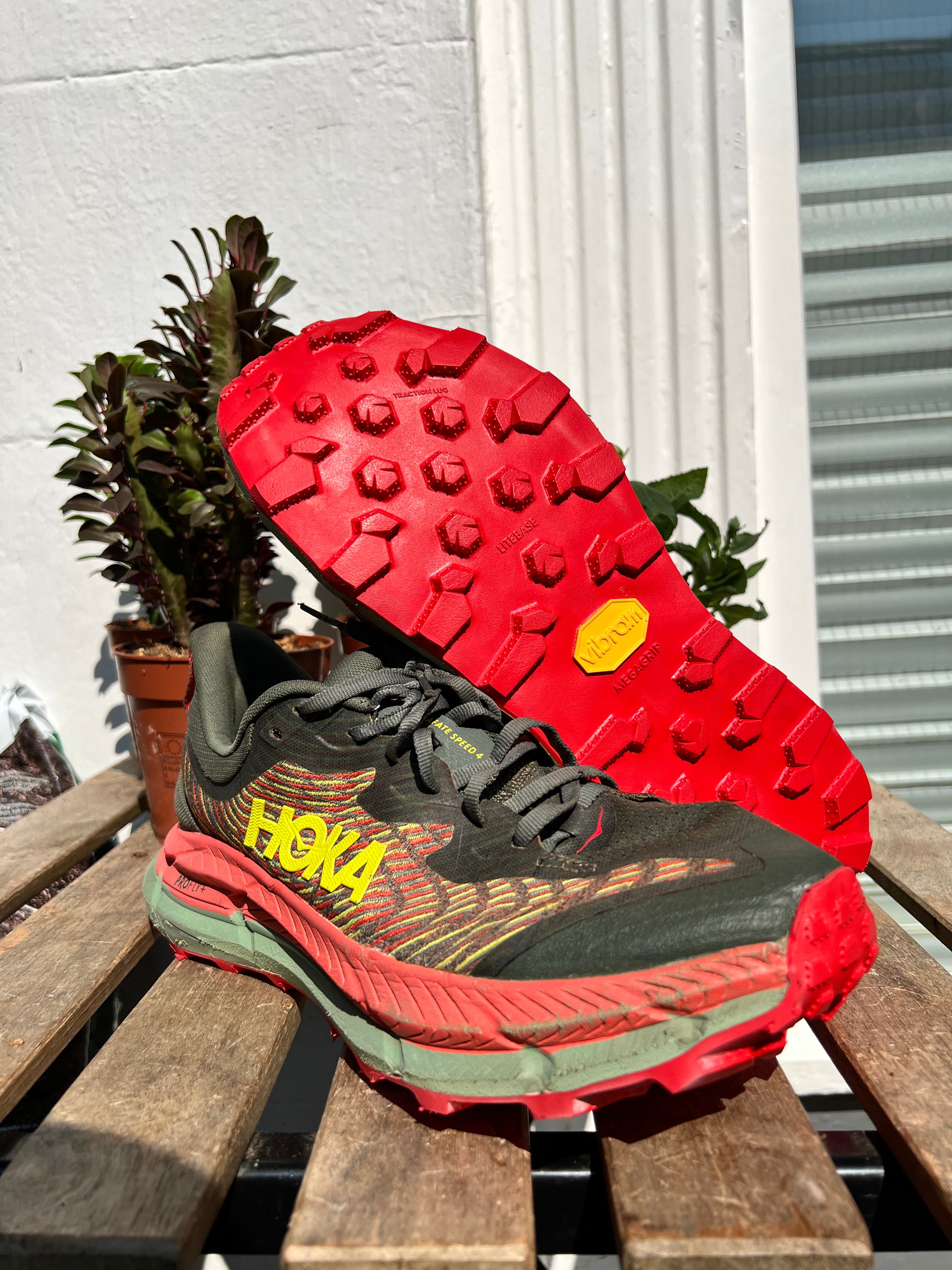 A pair of Hoka trail running shoes displayed on a wooden bench, with one shoe standing upright to show the side and the other lying on its side to show the red outsole. Running/Trail Resole (extended sizes 49-51) - Sole and Trail