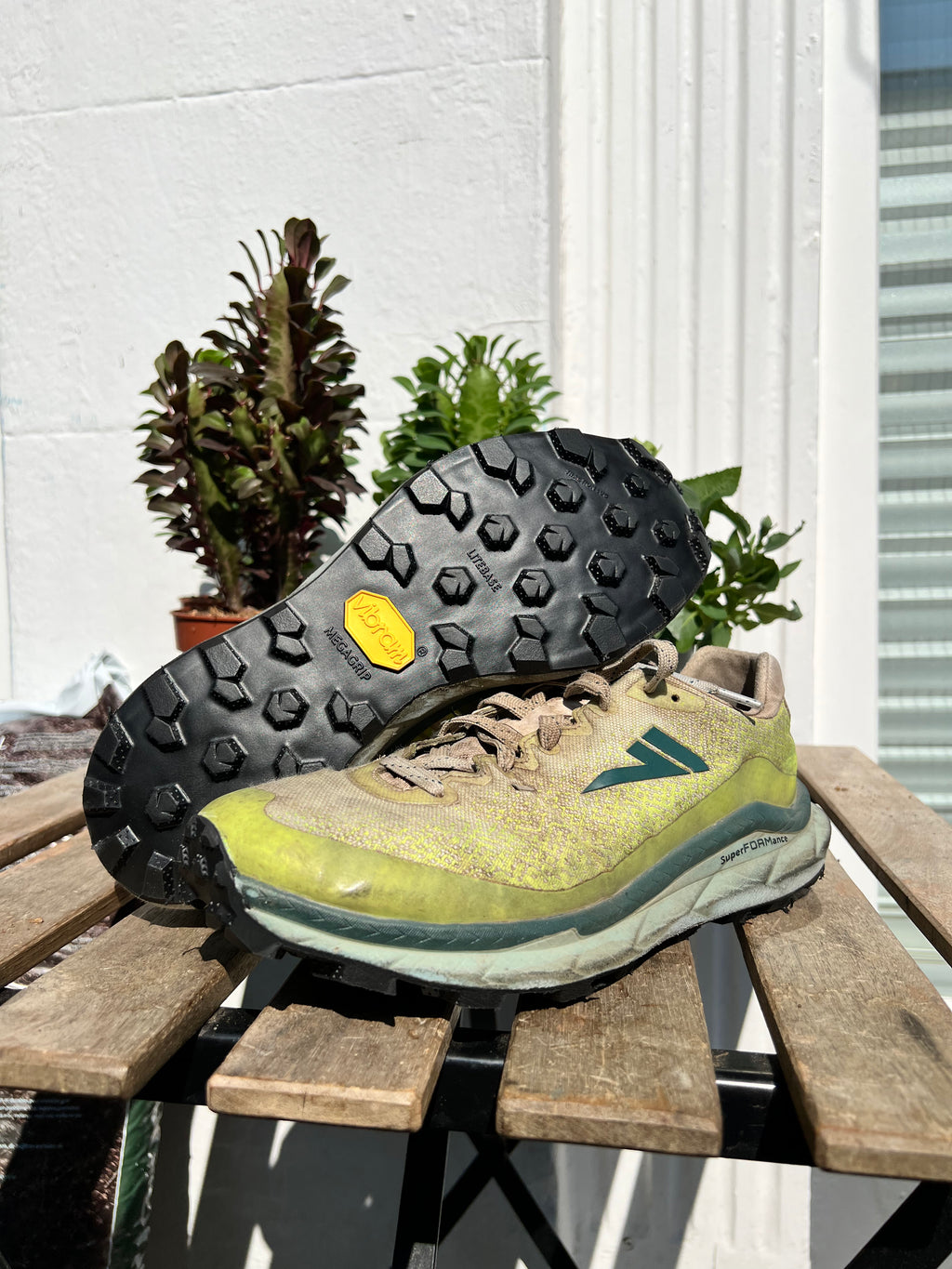 A pair of trail running shoes resting on a wooden table outdoors, with one shoe turned upside down to show the black treaded outsole and a yellow Vibram logo. The shoes have a yellow-green and gray suede/mesh upper with a teal logo. Running/Trail Resole (extended sizes 49-51) - Sole and Trail