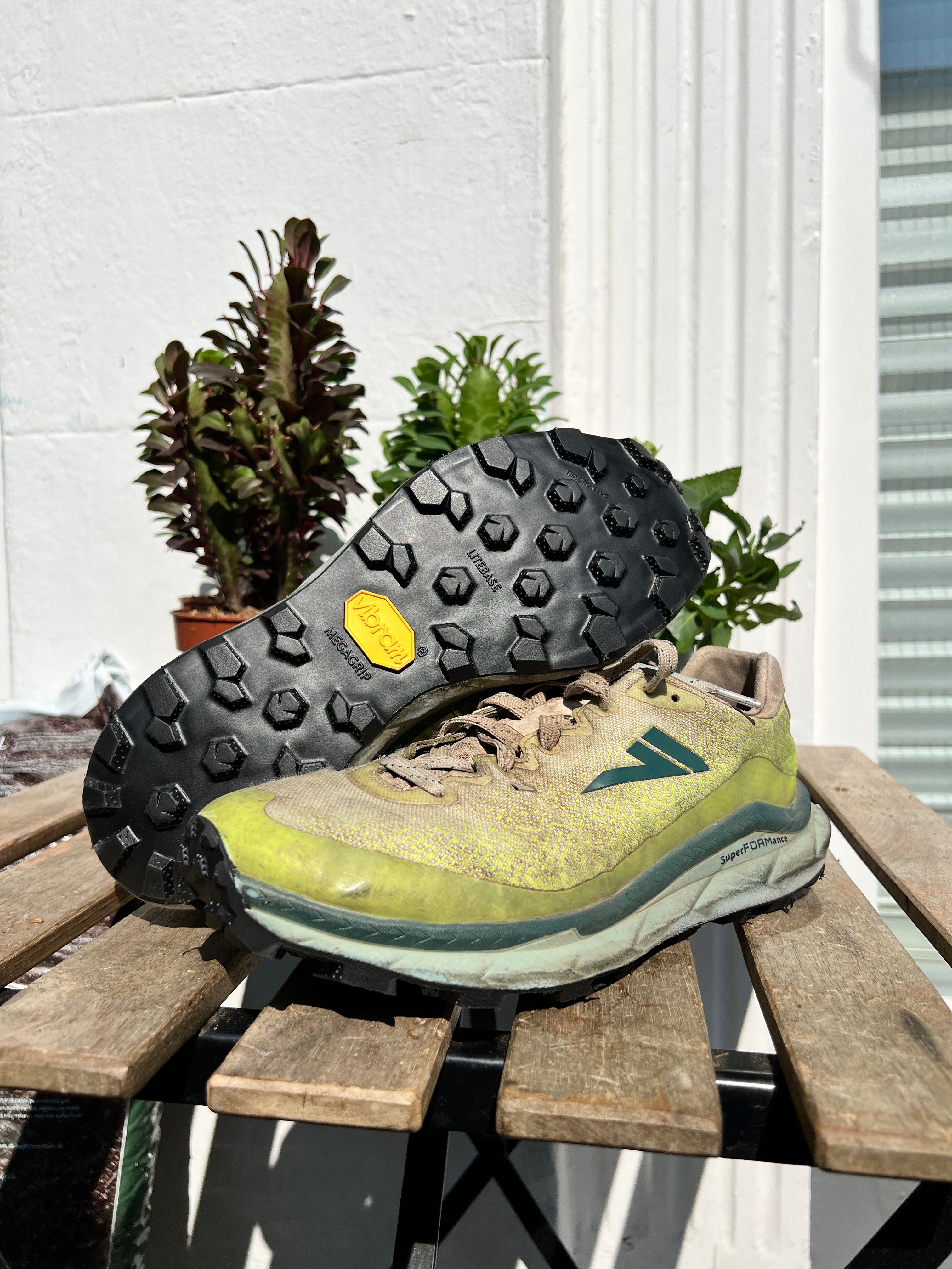 A pair of trail running shoes resting on a wooden table outdoors, with one shoe turned upside down to show the black treaded outsole and a yellow Vibram logo. The shoes have a yellow-green and gray suede/mesh upper with a teal logo. Running/Trail Resole (extended sizes 49-51) - Sole and Trail