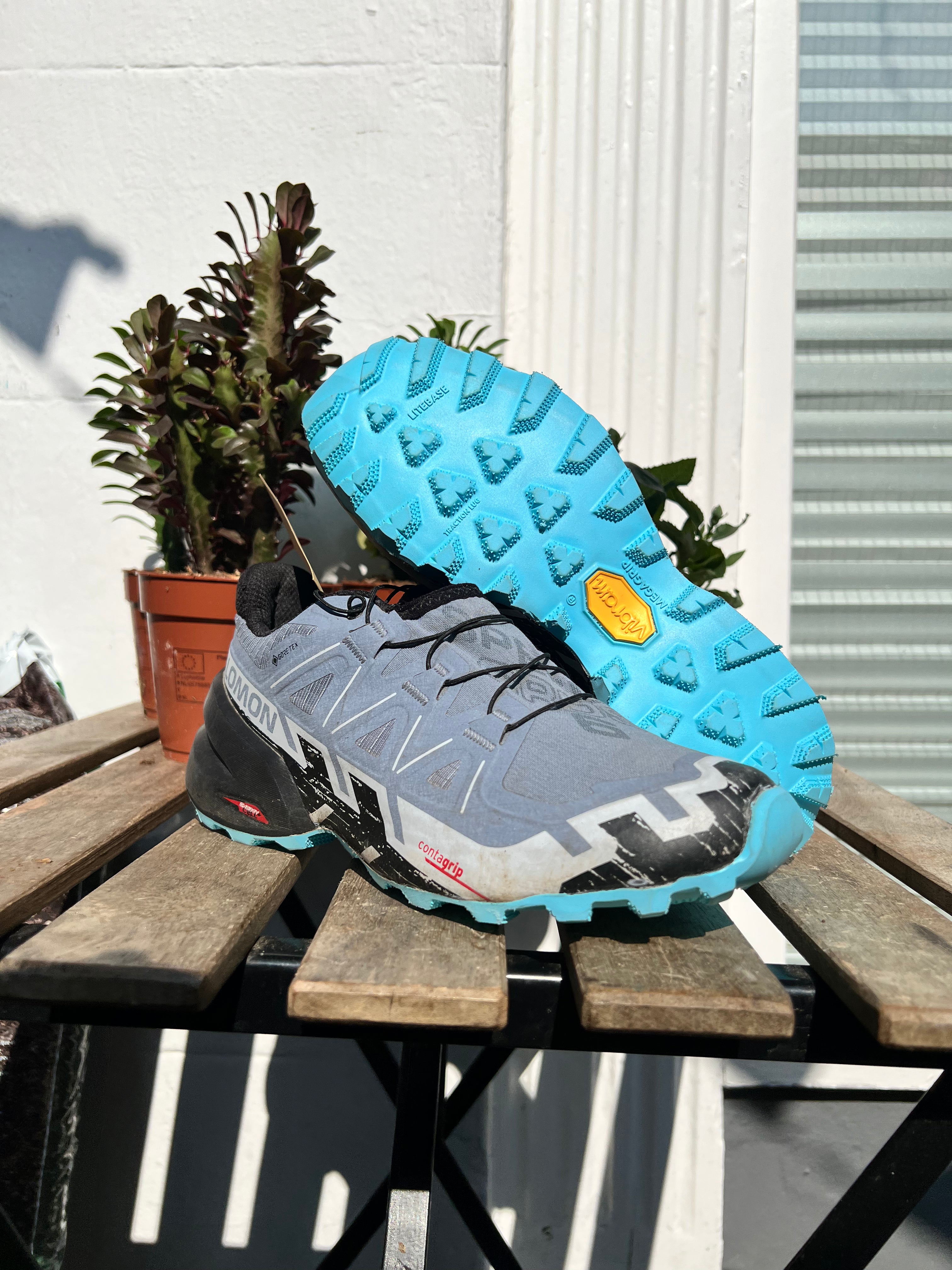 A pair of Salomon trail running shoes in gray, black, and bright blue, with one shoe showing the side profile and the other showing the textured blue outsole, displayed on a wooden table with potted plants in the background. Running/Trail Resole (extended sizes 49-51) - Sole and Trail