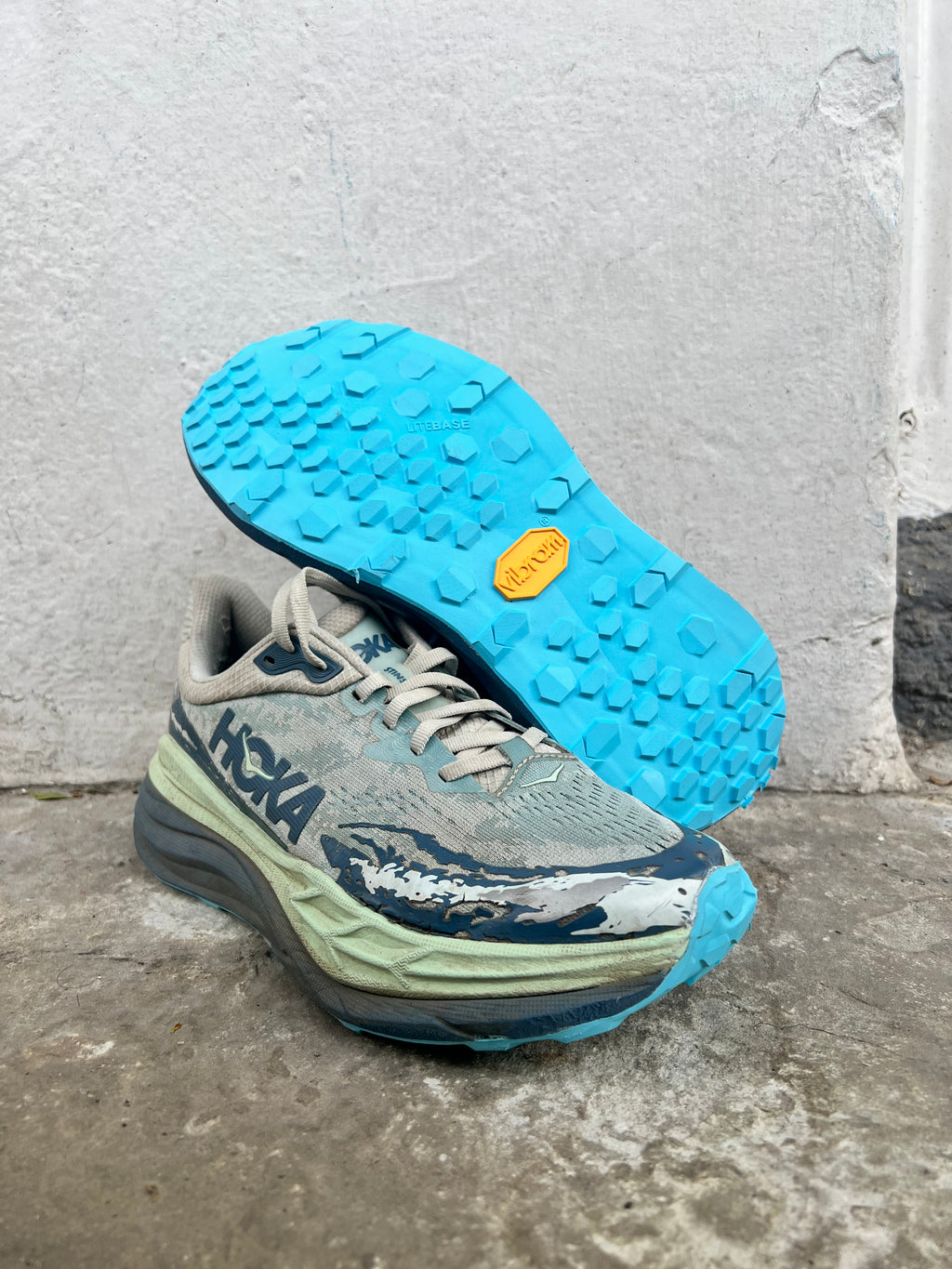 A pair of HOKA trail running shoes in a worn, used condition, resting on a concrete floor against a textured white wall. One shoe is upright showing the side with a blue HOKA logo, while the other lies on its side displaying a bright blue Vibram sole.