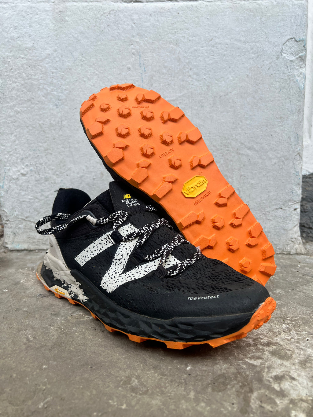 A pair of New Balance trail running shoes in black and orange, with one shoe standing upright to show the side profile and the other lying flat to display the textured orange outsole. The shoes are positioned on a concrete floor against a light grey concrete wall. Running/Trail Resole - Sole and Trail
Vibram zegatarc