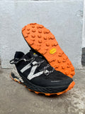 A pair of New Balance trail running shoes in black and orange, with one shoe standing upright to show the side profile and the other lying flat to display the textured orange outsole. The shoes are positioned on a concrete floor against a light grey concrete wall. Running/Trail Resole - Sole and Trail
Vibram zegatarc