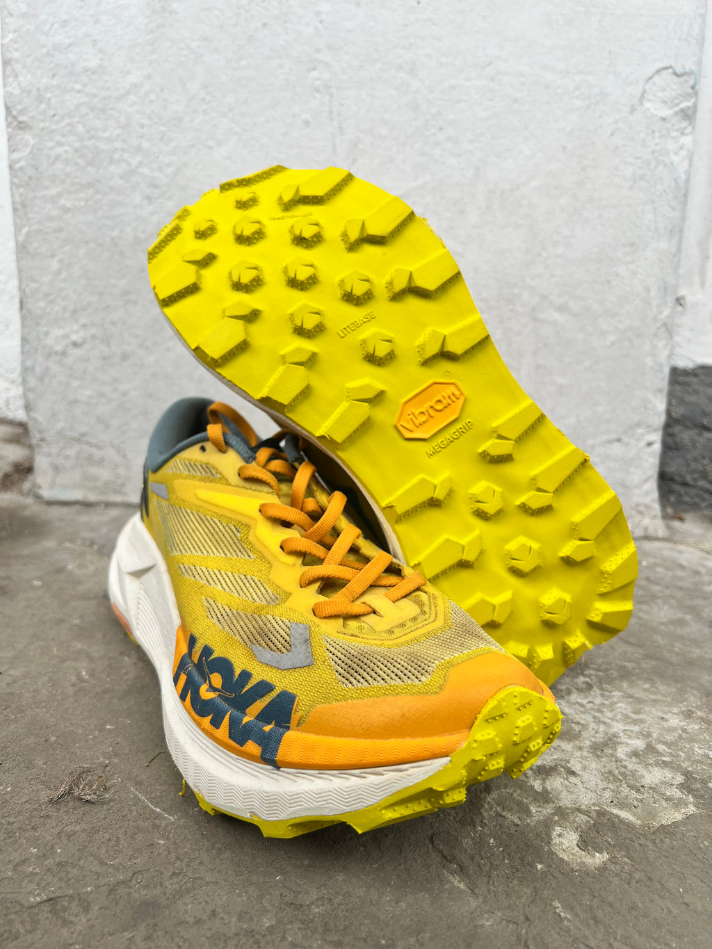 A pair of Hoka trail running shoes in yellow, orange, and white, with one shoe standing upright to show the textured yellow outsole and the other lying on its side showing the side profile with the HOKA logo. Vibram Zegatrac resole