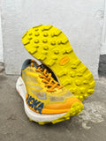 A pair of Hoka trail running shoes in yellow, orange, and white, with one shoe standing upright to show the textured yellow outsole and the other lying on its side showing the side profile with the HOKA logo. Vibram Zegatrac resole