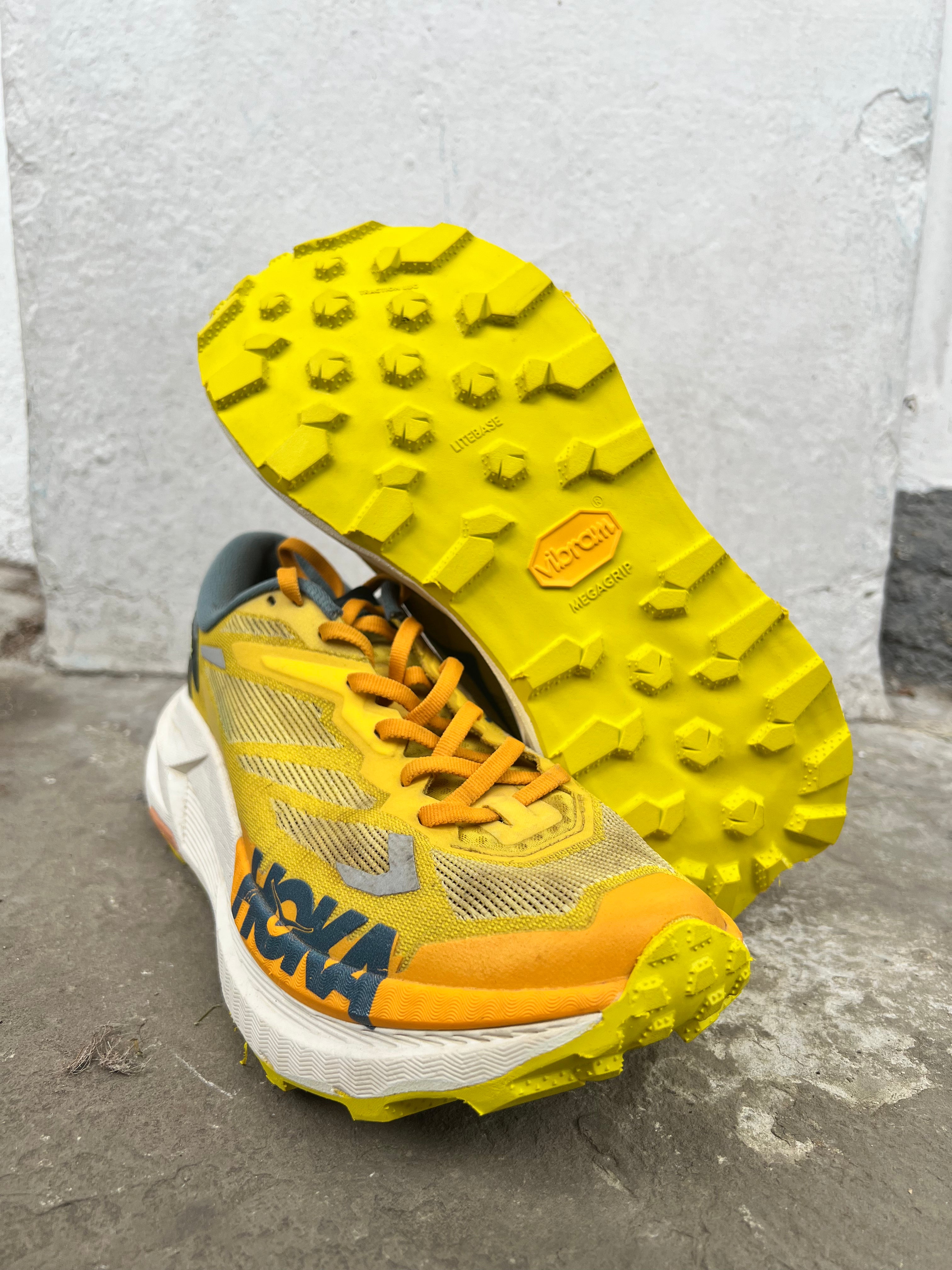 A pair of Hoka trail running shoes in yellow, orange, and white, with one shoe standing upright to show the textured yellow outsole and the other lying on its side showing the side profile with the HOKA logo. Vibram Zegatrac resole