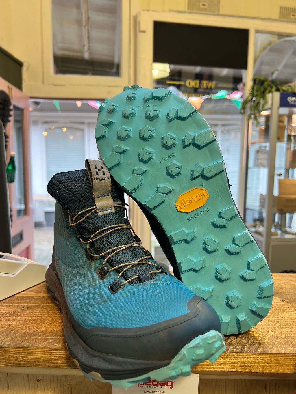 A pair of Vibram mid-height hiking boots in blue and dark gray, with one boot standing upright and the other tilted to show the textured teal outsole. The boots are displayed on a wooden counter inside a shop.Running/Trail Resole (extended sizes 49-51) - Sole and Trail