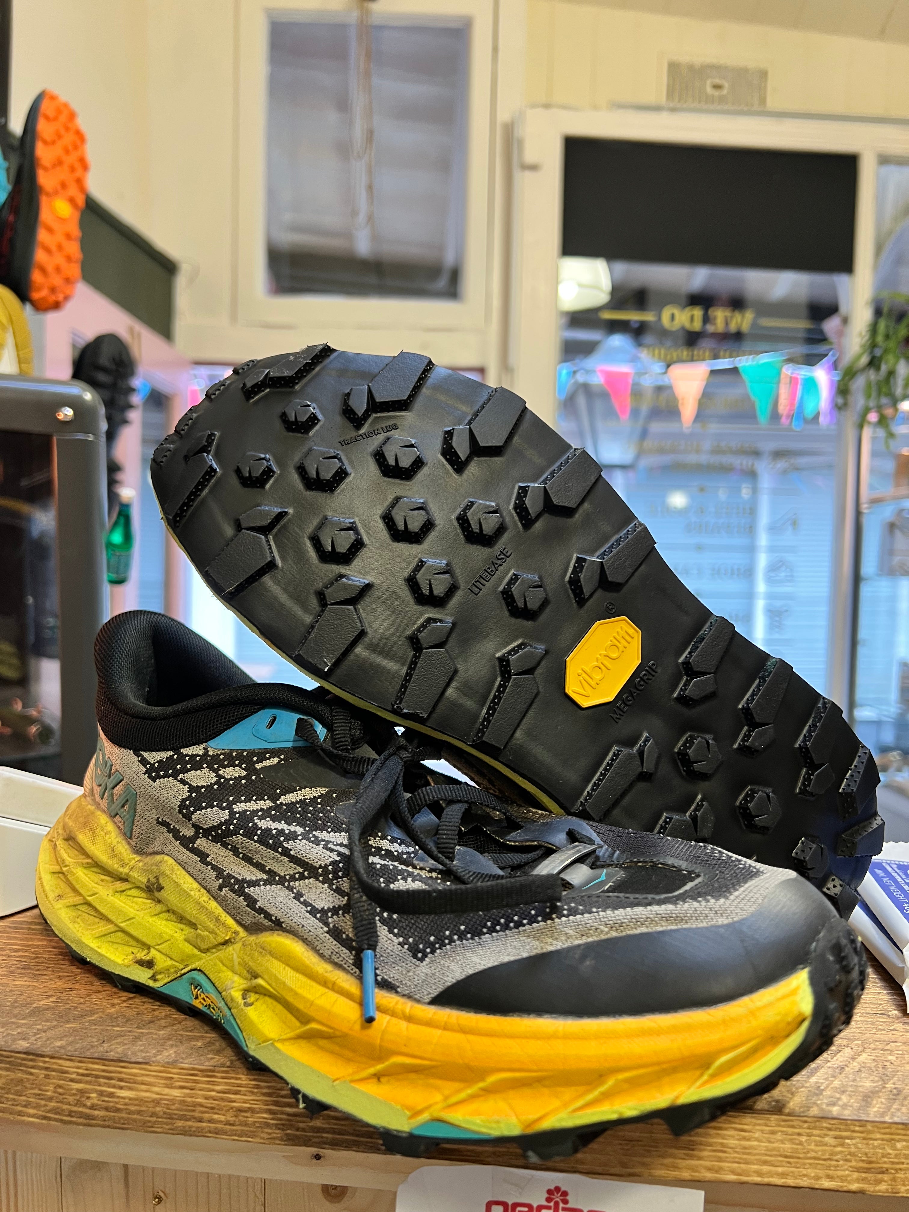 A pair of trail running shoes displayed on a wooden shelf in a store. One shoe is positioned upright to show the side profile, while the other is turned upside down to display the deep lugs of the outsole and the Vibram logo on the heel. Running/Trail Resole (extended sizes 49-51) - Sole and Trail
