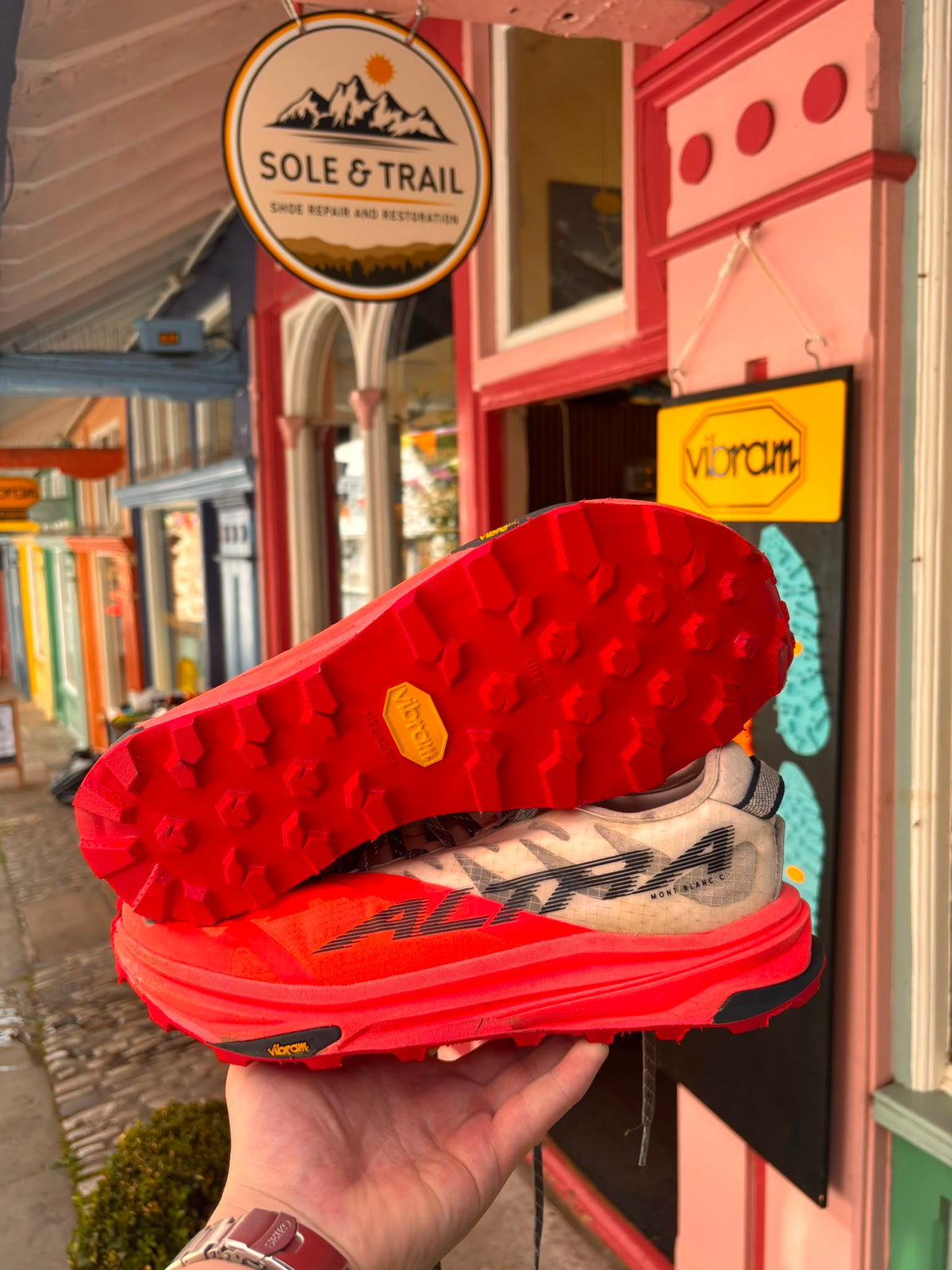 A hand holds a pair of red and gray Altra trail running shoes, showing the textured red Vibram sole and the side of the shoe with 'ALTRA' branding, in front of a storefront with a 'Sole & Trail' sign. Running/Trail Resole - Sole and Trail
Vibram zegatarc