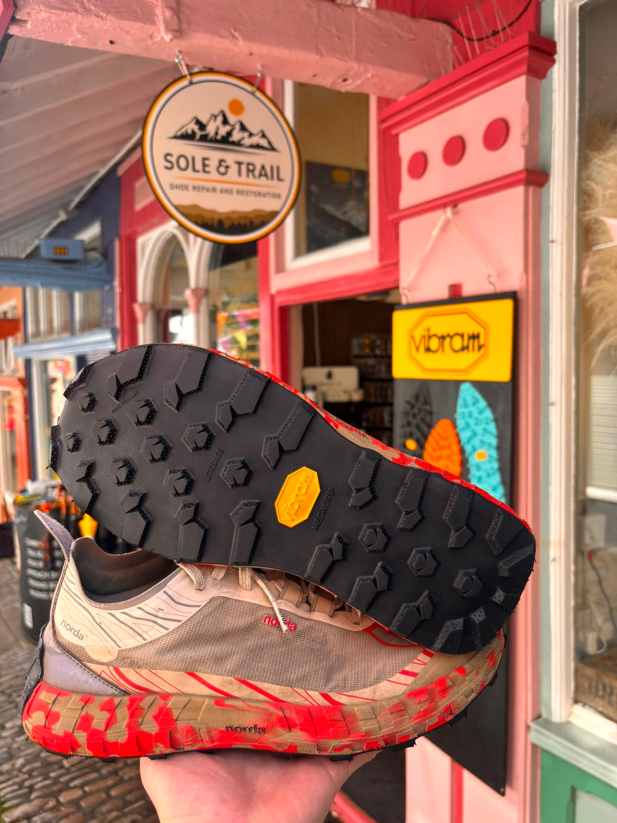 A person holds up the outsole of a trail running shoe to show the tread pattern and Vibram logo, with the shoe resting underneath. The shoe has a beige mesh upper with red and orange accents. Running/Trail Resole - Sole and Trail
Vibram zegatarc